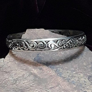 May include: A silver-colored bangle bracelet with an intricate, swirling, and dotted pattern. The bracelet is displayed on a gray, textured surface against a dark background. The design features a repeating scroll motif.