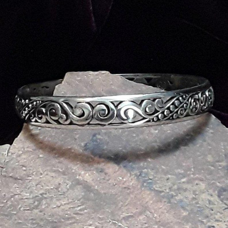 May include: A silver-colored bangle bracelet with an intricate, swirling, and dotted pattern. The bracelet is displayed on a gray, textured surface against a dark background. The design features a repeating scroll motif.