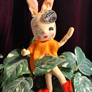 May include: A vintage handmade Mop-Pet doll, featuring a bunny-like face with long ears, rosy cheeks, and red lips. The doll wears an orange dress and red shoes. A tag reads "Handmade Imported Mop-Pets". The doll is posed among green leaves.