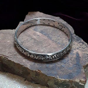 May include: A silver-colored bangle bracelet with an intricate, swirling pattern. The bracelet is circular and rests on a rough, textured stone surface. The design includes floral and scroll-like elements.
