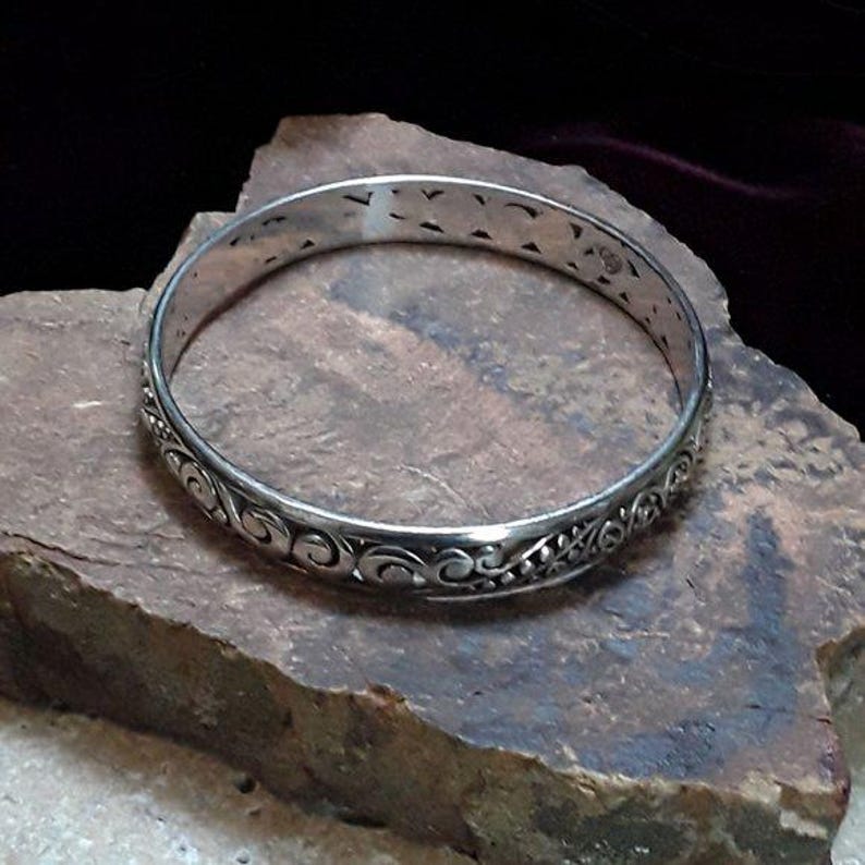 May include: A silver-colored bangle bracelet with an intricate, swirling pattern. The bracelet is circular and rests on a rough, textured stone surface. The design includes floral and scroll-like elements.