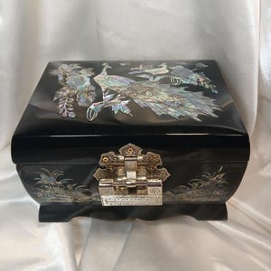 May include: A rectangular black lacquered jewelry box featuring iridescent mother-of-pearl inlay depicting a peacock and floral motifs on the lid and front. The box includes a silver-toned metal latch closure and four small feet.