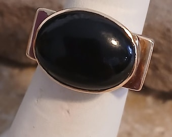 May include: A gold ring with a large, oval-shaped black gemstone. The ring has a simple, modern design.