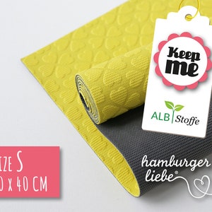 Könnte beinhalten: Eine gelbe und graue Yogamatte mit einem Herzmuster. Die Matte ist aufgerollt und hat ein Etikett mit der Aufschrift "Keep me" und "ALB Stoffe". Die Matte ist mit "Größe S 30 x 40 CM" und "hamburger liebe" gekennzeichnet.