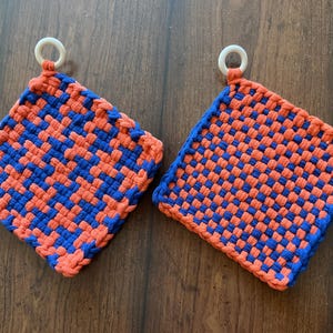 May include: Two woven potholders in a basket weave pattern. The potholders are orange and blue, with a white ring for hanging. The potholders are square and have a thick, textured appearance. The potholders are on a dark wooden surface.