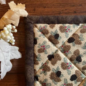 May include: A quilted square with a brown border and a beige center patterned with pinecones, green pine needles, and red berries. The corner of the quilt is visible, showcasing the detailed stitching and fabric design. Decorative elements are in the upper left corner.