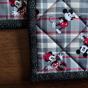 May include: Close-up of a quilted potholder featuring a gray and white plaid pattern with red accents. The potholder is decorated with images of Minnie Mouse in her signature red polka-dot dress and bow. The border is a dark gray with white polka dots.