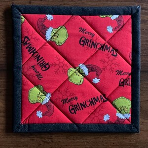 May include: Square quilted potholder with a red background featuring the Grinch in a Santa hat and the words "Merry Grinchmas." The potholder has a black border and a diamond pattern.
