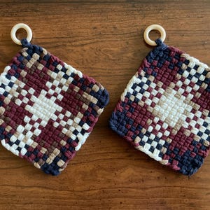 May include: Two woven potholders with a geometric pattern in white, burgundy, navy blue, and tan. Each square potholder has a small, off-white ring attached for hanging. The potholders are displayed on a wooden surface.