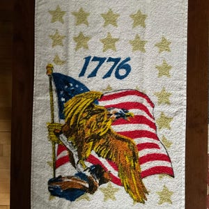 May include: White rectangular towel with a fringed edge, featuring a patriotic design. The design includes a waving American flag, a golden eagle, gold stars, and the blue text "1776".