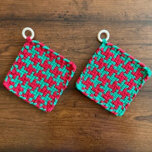 May include: Two handmade, square pot holders with a woven pattern in red and green. Each pot holder has a white loop for hanging. The pot holders are displayed on a wooden surface.