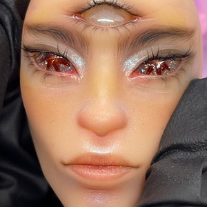 May include: A close-up of a person's face with three eyes. The eyes are surrounded by glitter and the person has a neutral expression.