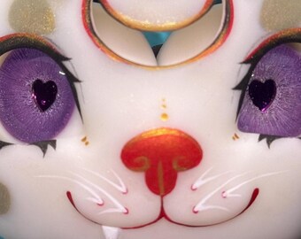 May include: A close-up of a white cat-like mask with large purple eyes, each with a heart-shaped pupil. The mask has a red nose, gold accents, and black eyelashes. The overall design is whimsical and artistic.