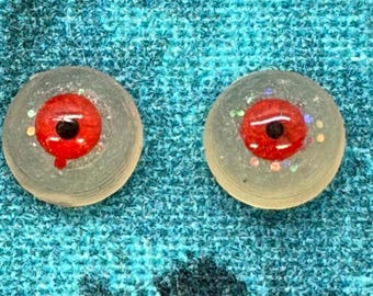 May include: Two circular, translucent resin cabochons featuring a realistic red iris and a black pupil. The clear base of each piece is embedded with small, iridescent glitter particles that catch the light, creating a subtle, shimmering effect.