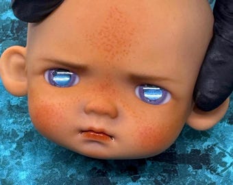 May include: A doll head with a fair complexion, freckles, and rosy cheeks. The doll has blue eyes with a reflective quality, a small nose, and a slightly downturned mouth. The head is held by a gloved hand.