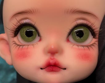 May include: Close-up of a doll's face with detailed features. The doll has large, green eyes with long eyelashes, delicate eyebrows, and rosy cheeks. The lips are painted a deep red, and the skin tone is pale.