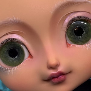 May include: Close-up of a doll's face with green eyes and delicate features. The doll has pale skin, pink eyeshadow, and light pink lips. White eyelashes and eyebrows add detail to the face.