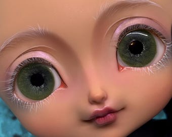 May include: Close-up of a doll's face with green eyes and delicate features. The doll has pale skin, pink eyeshadow, and light pink lips. White eyelashes and eyebrows add detail to the face.