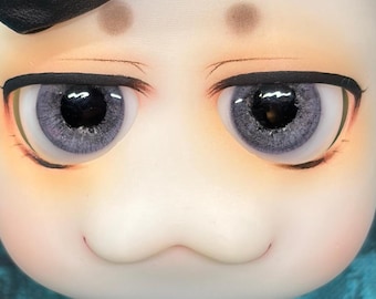 May include: A doll head featuring large, round, light purple eyes with detailed black pupils and irises. The face has a smooth, pale complexion with soft orange blush on the cheeks, two small brown spots above the eyes, and a subtle, thin mouth line.
