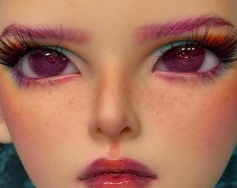 May include: Close-up of a doll's face with purple eyes, pink eyebrows, and long eyelashes. The doll has a fair complexion with freckles and glossy, dark red lips. The eye makeup includes teal eyeliner and pink eyeshadow.