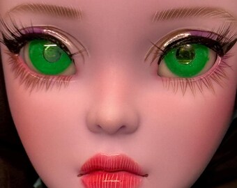 May include: Close-up of a doll's face with striking features. The doll has bright green eyes with long, black eyelashes, and full red lips. The skin is a pale pink, and the eyebrows are light brown. The doll's face is smooth and detailed.