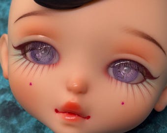 May include: Close-up of a doll's face with detailed makeup. The doll has large, purple eyes with long eyelashes, arched eyebrows, and a small, red mouth. Two small, pink dots are on the cheeks. The skin tone is pale with subtle blush.