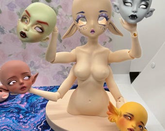 May include: A nude-colored art doll with multiple arms and a round base, holding several interchangeable doll heads. The heads are painted in various colors, including green, gray, and yellow, with detailed facial features and expressions.