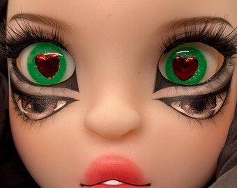 May include: Close-up of a doll face with green eyes featuring a red heart design. The doll has long, black eyelashes, detailed eye makeup, and red lipstick. The skin tone is pale, and the overall aesthetic is stylized.