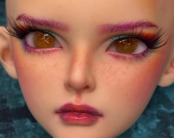 May include: Close-up of a doll face with detailed makeup. The doll has brown eyes, long black eyelashes, and purple eyebrows. The cheeks are blushed pink, and the lips are a glossy red. Freckles are visible on the face.