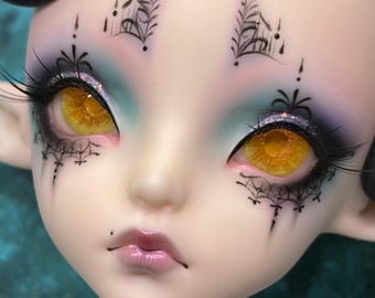 May include: A doll head with pale skin, bright yellow eyes, and long black eyelashes. The face features intricate black line art resembling spiderwebs around the eyes and forehead, with soft blue and purple eyeshadow and glossy pink lips.