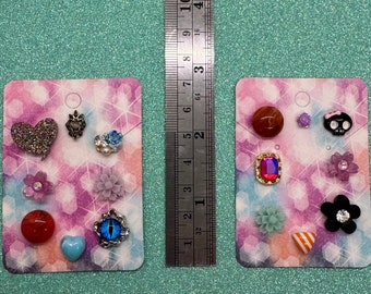 May include: Two cards with a variety of colorful earrings. The earrings are on a white background with a pink and purple pattern. The earrings include a heart, a skull, a flower, a star, an eye, a red gem, a blue gem, and a black gem.