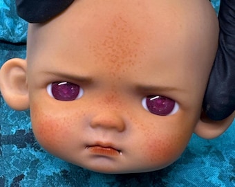 May include: A doll head with a neutral expression, featuring a light brown complexion, freckles, and rosy cheeks. The doll has large, vibrant purple eyes and a small, detailed mouth. The head is held by a gloved hand.
