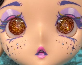 May include: Close-up of a doll's face with large, glittery brown eyes. The doll has purple and blue eye makeup, freckles, and a pink mouth. The overall aesthetic is whimsical and artistic.
