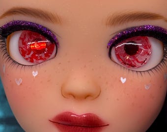 May include: Close-up of a doll's face with striking features. The doll has red eyes with intricate details, framed by purple glitter eyeshadow and long lashes. White heart-shaped accents are on the cheeks, and the lips are a deep red.