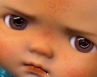 May include: Close-up of a doll's face with a slightly furrowed brow. The doll has brown eyes with a reflective quality, rosy cheeks, and freckles. The lips are glossy and slightly parted, giving a subtle expression.