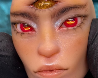 May include: Close-up of a face with red eyes and a third eye. The eyes have a reflective, fiery appearance. The skin is smooth with subtle highlights. The third eye is gold with glitter. The eyebrows are dark and well-defined.
