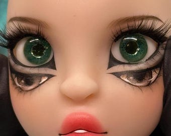 May include: Close-up of a doll's face with striking features. The doll has large, green eyes with long, black eyelashes and dramatic black eyeliner. The lips are painted a bright red, and the skin is pale. The overall aesthetic is stylized and artistic.