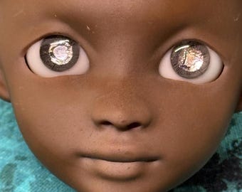 May include: Close-up of a doll's face with dark brown skin. The doll has large, reflective eyes with a detailed, multi-colored iris. The face is smooth with subtle details around the nose and mouth, and the overall expression is neutral.