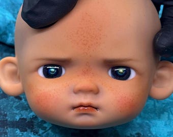 May include: A close-up of a doll's head with a fair complexion, rosy cheeks, and freckles. The doll has large, reflective blue eyes and a slightly pursed mouth. The doll's head is being held by a gloved hand.