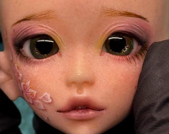 May include: Close-up of a doll's face with detailed makeup. The doll has green eyes, pink and purple eyeshadow, and delicate pink cherry blossom designs on the cheek. The doll's lips are a natural pink color.