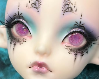 May include: A doll face with pale skin, bright pink irises, and long black eyelashes. The eyes are framed by intricate black line art resembling spiderwebs and decorative patterns. The eyelids feature blue and purple eyeshadow, and the lips are pink.
