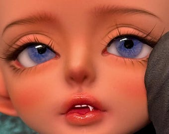 May include: Close-up of a doll's face with detailed features. The doll has light skin, rosy cheeks, and full lips. The eyes are a striking blue with long, dark eyelashes. The doll's expression is one of slight surprise.