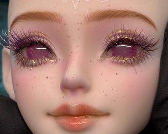 May include: A doll face featuring pale skin with brown freckles, light brown eyebrows, and vibrant pink eyes. The eyes are framed by long, dark purple eyelashes and accented with gold glitter along the lower lash lines. The lips are a glossy peach tone.