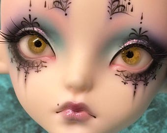 May include: Close-up of a doll's face with detailed makeup. The doll has large, golden eyes with long eyelashes and intricate black designs around the eyes. The skin is pale, and the lips are a glossy pink.