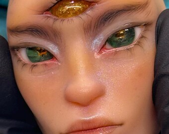 May include: Close-up of a face with three eyes. The two lower eyes are green, and the upper eye is gold. The skin has a smooth texture with subtle highlights. The eyebrows are dark, and the lips are a natural color.