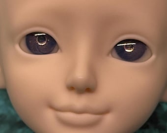May include: Close-up of a doll's face with a smooth, pale complexion. The doll has large, expressive eyes with a deep purple iris and reflective highlights. The nose is small and the lips are gently curved, creating a serene expression.