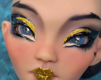 May include: A doll head featuring bright blue eyes with heavy black eyeliner and gold glitter accents on the eyelids. The lips are painted with a textured gold glitter finish, and a small black heart is painted on the left cheek near the eye.
