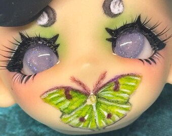 May include: Close-up of a doll face with lavender eyes and long black eyelashes. A green and yellow butterfly is painted on the doll's face. The doll has a pale complexion with pink blush.