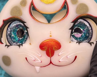 May include: Close-up of a white, round, cat-like face with large, glittery blue-green eyes. The nose is orange, and the mouth is a red smile with a single fang. Gold accents and a crescent moon shape are above the eyes.