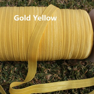 May include: A spool of gold yellow elastic fold over elastic, also known as FOE, is shown on a white cardboard spool. The elastic is wound tightly and is ready to be used for sewing projects.